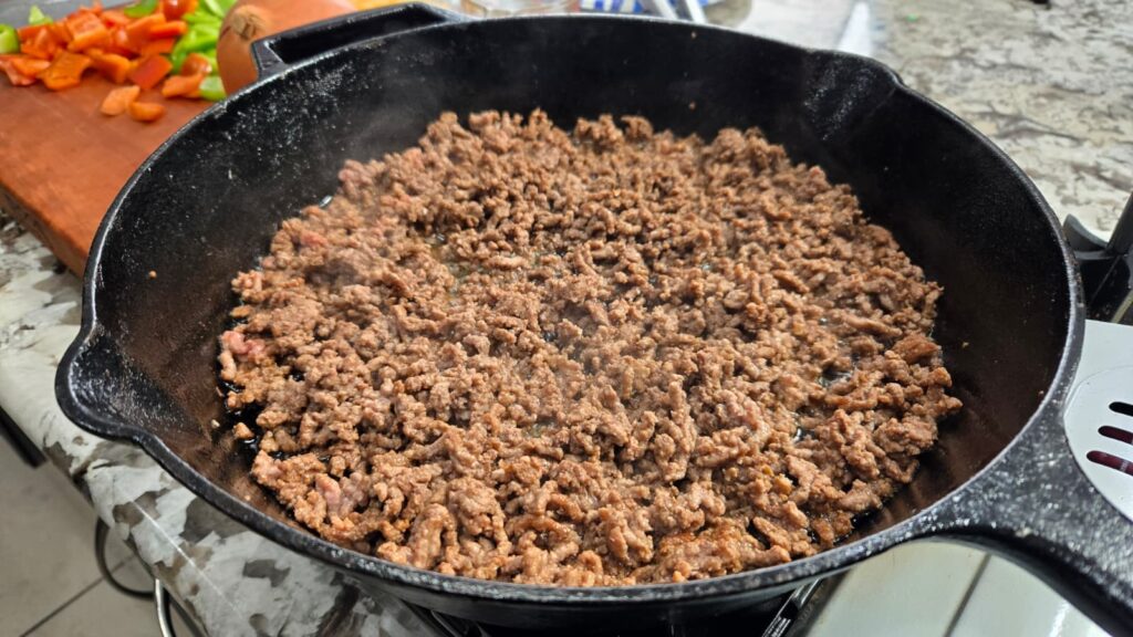 A cast iron cornbread skillet filled with browned ground beef cooking on a stovetop, with steam rising. Chopped bell peppers and a knife are visible on a cutting board in the background, hinting at a hearty dirty cabbage meal in progress.