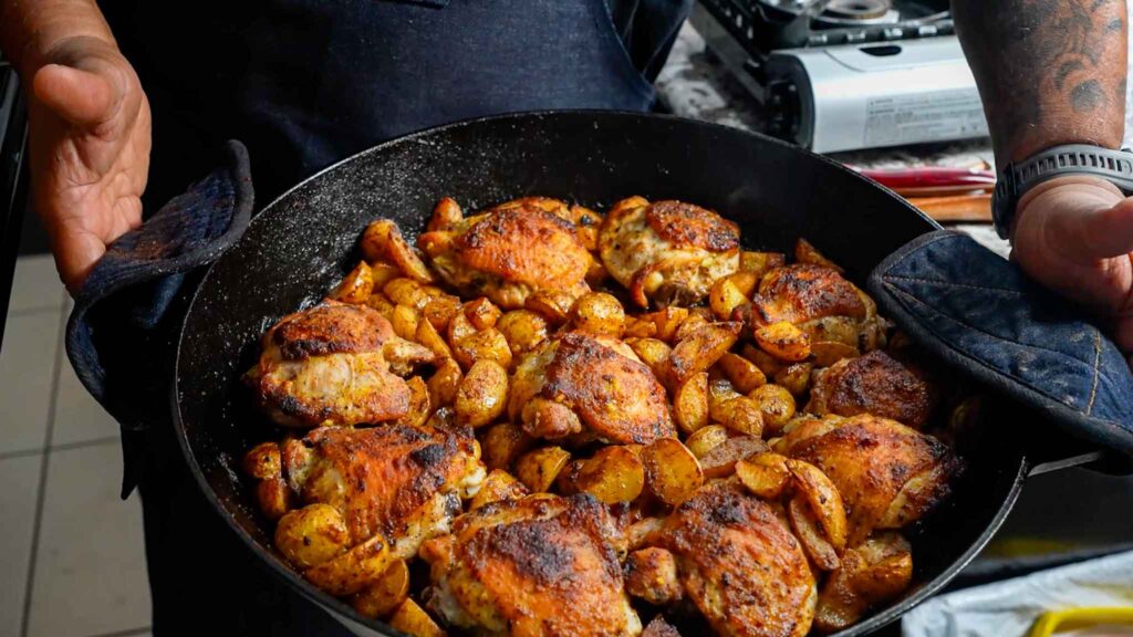 A person holding a black skillet filled with honey mustard chicken thighs and seasoned potatoes, fresh from the oven, with a stove in the background.