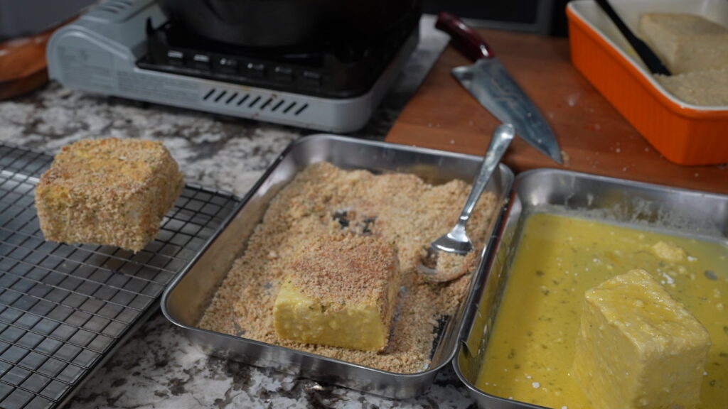 A breaded block, perfect for making fried grit cakes and shrimp, sits on a rack, while another block is coated in breadcrumbs in a tray next to a fork and an egg wash tray. A knife and baking dish are visible in the background.