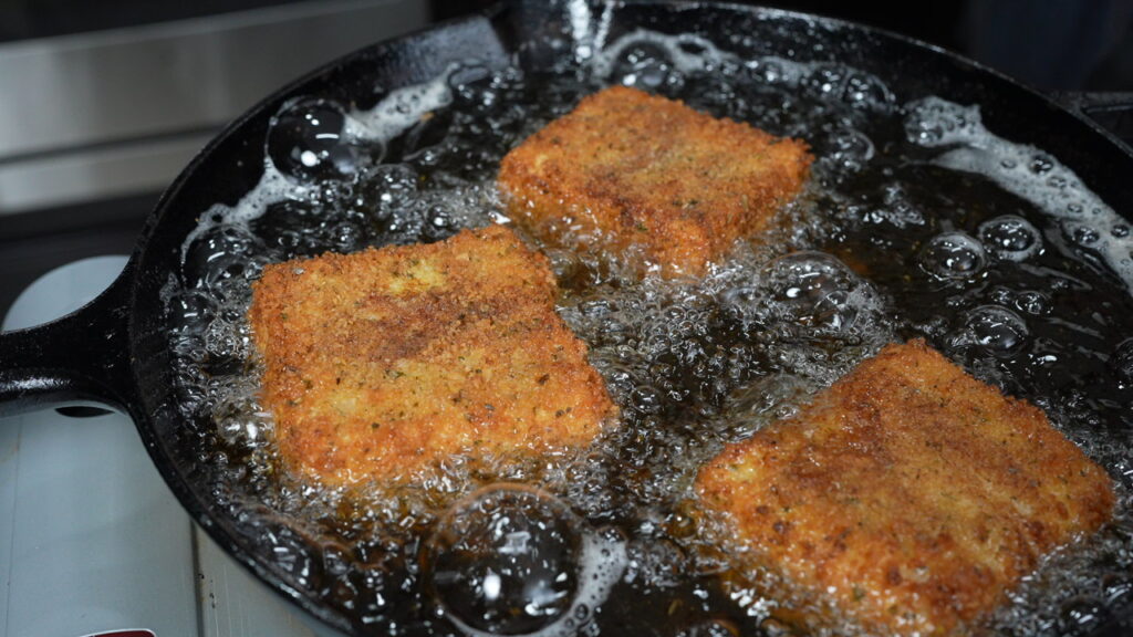 Three breaded squares of fried grit cakes are frying in hot oil inside a black cast iron skillet, with bubbles forming around the food as it cooks&mdash;perfectly crisp and ready to be paired with savory shrimp.