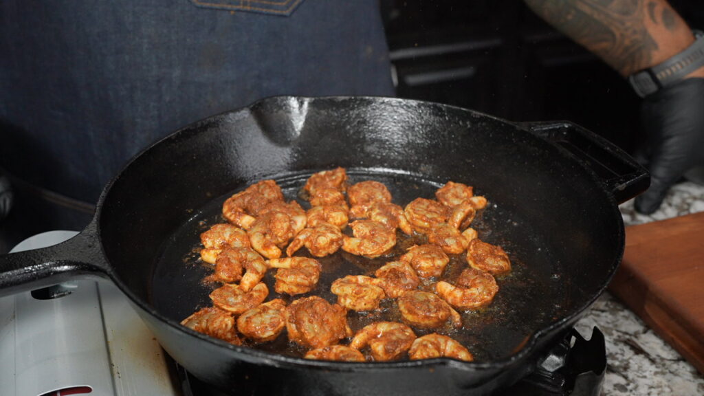 Seasoned shrimp cooking in a black cast iron skillet on a stove, with a person in a dark apron and gloves standing nearby. On the right side, a wooden cutting board sits ready to serve up crispy fried grit cakes and shrimp.