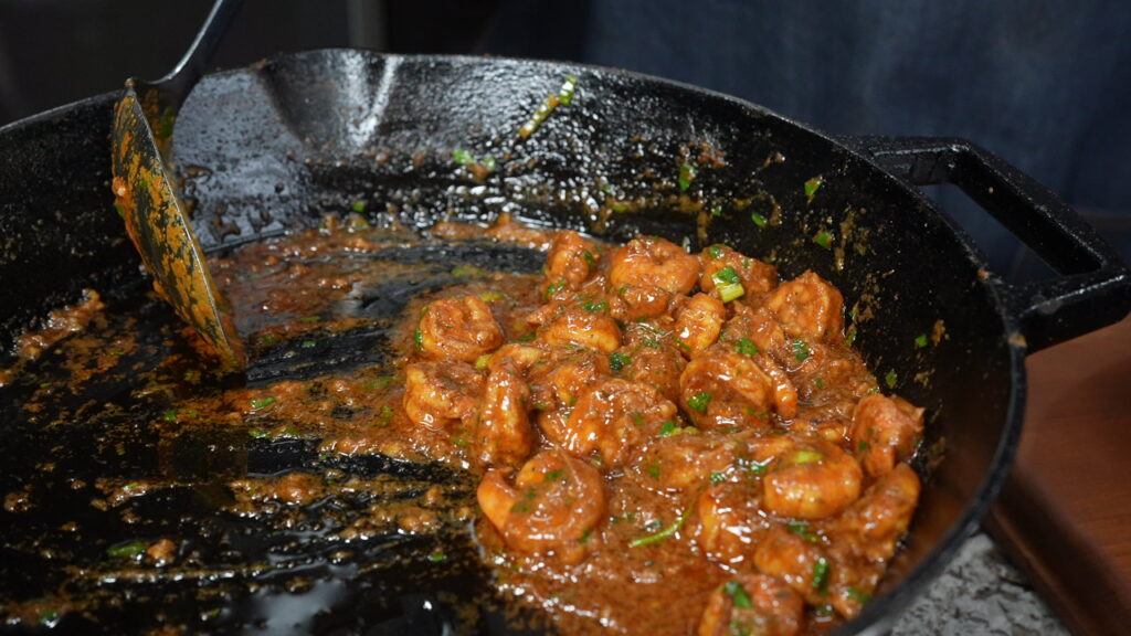 Shrimp cooking in a cast iron skillet with a thick, reddish-brown sauce and green herbs, perfect as a topping for fried grit cakes and shrimp. A metal spatula rests on the side of the pan.