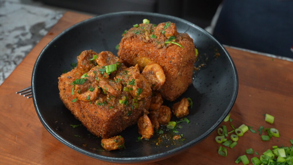 A black bowl holds two large fried grit cakes topped with saucy shrimp and garnished with chopped green onions and herbs, sitting on a wooden surface.