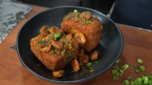 A black bowl holds two large fried grit cakes topped with saucy shrimp and garnished with chopped green onions and herbs, sitting on a wooden surface.