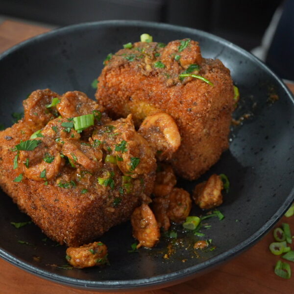A black bowl holds two large fried grit cakes topped with saucy shrimp and garnished with chopped green onions and herbs, sitting on a wooden surface.