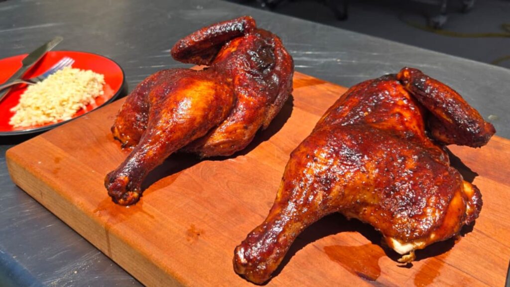 Two grilled bbq chicken halves, smoked and seasoned to perfection, rest on a wooden cutting board. In the background, a red plate with rice and a fork adds color to this inviting outdoor scene on a metal surface.