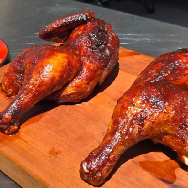 Two grilled bbq chicken halves, smoked and seasoned to perfection, rest on a wooden cutting board. In the background, a red plate with rice and a fork adds color to this inviting outdoor scene on a metal surface.