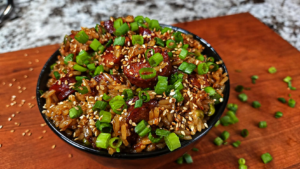 A black bowl filled with fried rice, topped with sliced sausage, chopped green onions, and sesame seeds, sits on a wooden surface with scattered green onions.