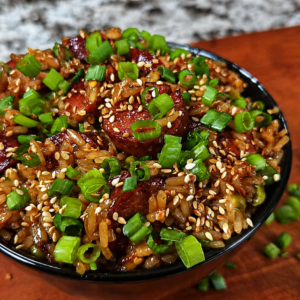 A black bowl filled with fried rice, topped with sliced sausage, chopped green onions, and sesame seeds, sits on a wooden surface with scattered green onions.