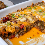 A cheesy ground beef casserole topped with melted cheese and chopped green onions, with a serving scooped out onto a plate in the background. Orange juices pool in the baking dish.