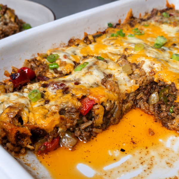 A cheesy ground beef casserole topped with melted cheese and chopped green onions, with a serving scooped out onto a plate in the background. Orange juices pool in the baking dish.