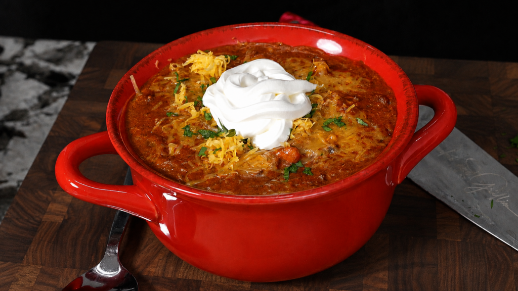 A red bowl filled with andouille chili, topped with shredded cheese, chopped herbs, and a dollop of sour cream, sits on a wooden surface with a spoon and knife nearby.