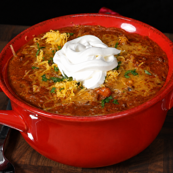 A red bowl filled with andouille chili, topped with shredded cheese, chopped herbs, and a dollop of sour cream, sits on a wooden surface with a spoon and knife nearby.