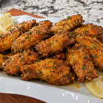 A white platter holds a dozen seasoned chicken wings garnished with herbs and lemon wedges, set on a marbled countertop.