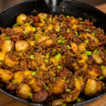 A cast iron skillet filled with dirty smothered potatoes, cooked ground beef, sliced sausage, halved baby potatoes, melted cheddar cheese, and chopped green peppers on a wooden table.