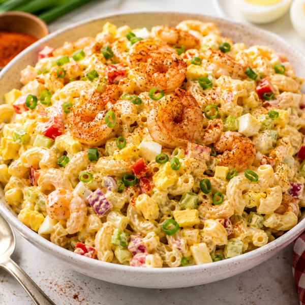 A bowl of creamy macaroni salad topped with seasoned shrimp, chopped green onions, diced red and yellow peppers, celery, and hard-boiled eggs, next to a spoon and a red gingham napkin.