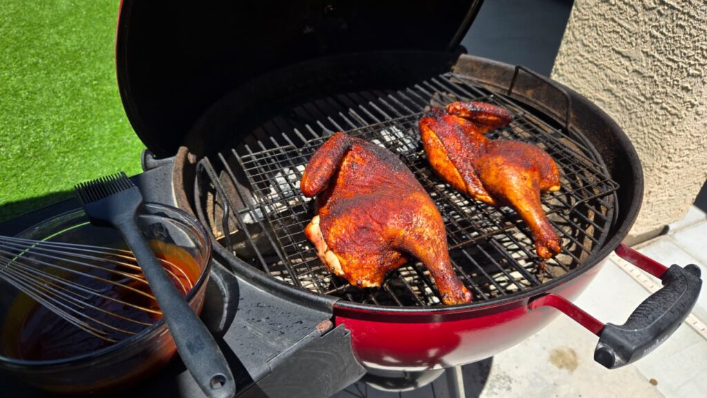 Two seasoned, grilled BBQ chicken halves are spatchcocked on a round charcoal barbecue. A bowl of barbecue sauce, basting brush, and whisk sit nearby, with green artificial grass and a wall in the background.