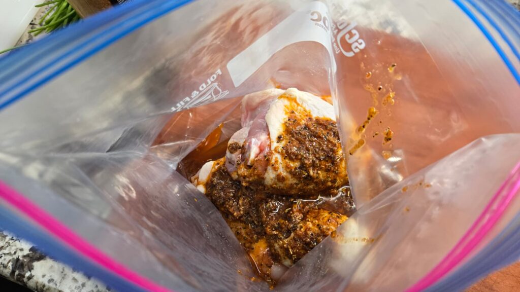 Raw chicken pieces coated in a dark spice marinade sit inside a clear resealable plastic bag, ready to be marinated. The bag is open and placed on a kitchen countertop.