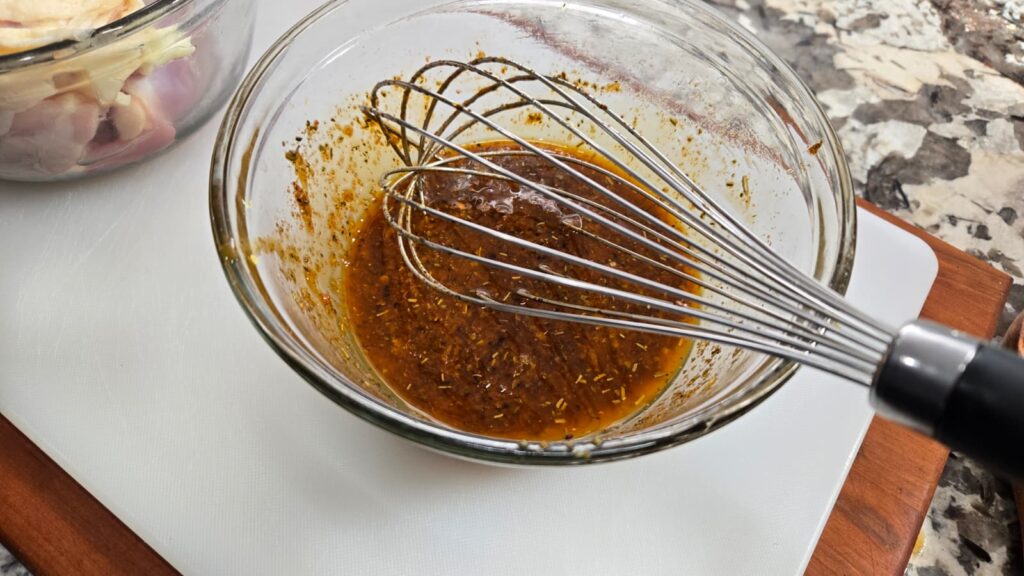 A glass bowl contains a reddish-brown marinade mixture being whisked with a metal whisk. In the background, there is a bowl of raw chicken pieces on a countertop.