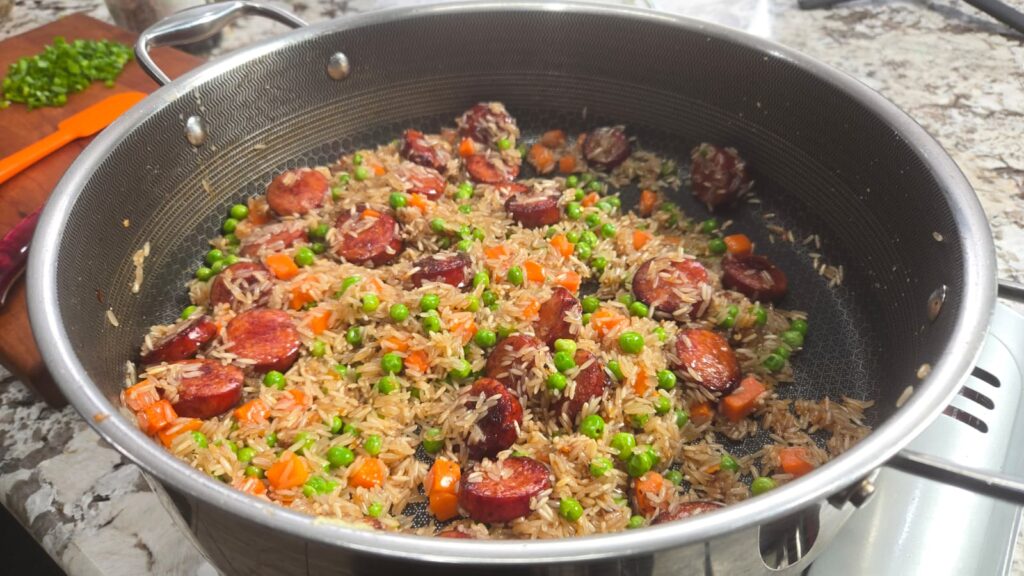 Rice with sliced sausage, peas, and diced carrots cooking in a stainless steel pot on a stovetop. Chopped green onions and an orange spatula are on a wooden cutting board in the background.