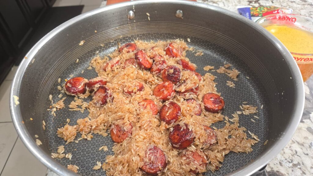 Brown rice and sliced sausage are being cooked in a nonstick pan. The sausage pieces are browned, and the rice looks slightly toasted. There is also a bowl with a yellow liquid mixture visible to the side.