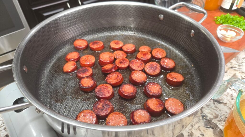 Sliced sausage pieces are sizzling and browning in a large metal pan on a stove. The sausages are evenly spread out and surrounded by bubbling oil.