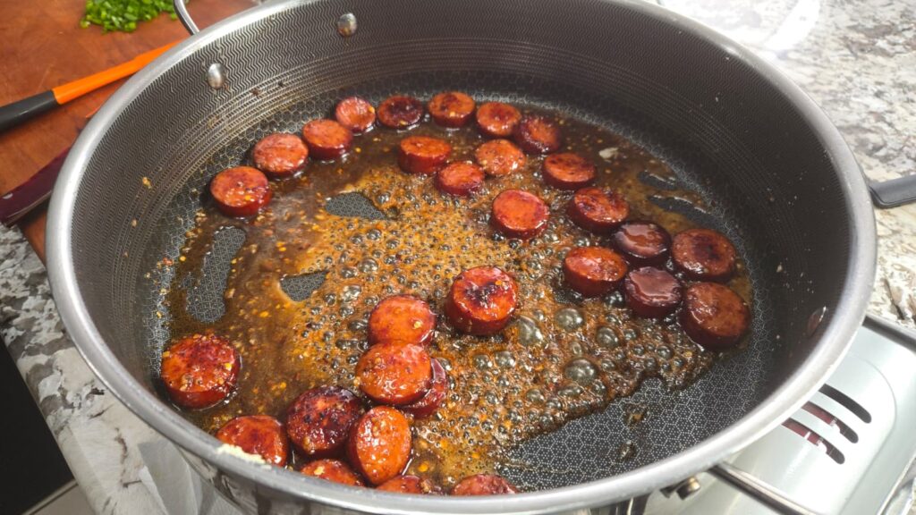 Sliced sausages are being cooked in a frying pan with oil and seasonings, creating a sizzling effect. The pan is on a stovetop, and there is a cutting board with a knife in the background.
