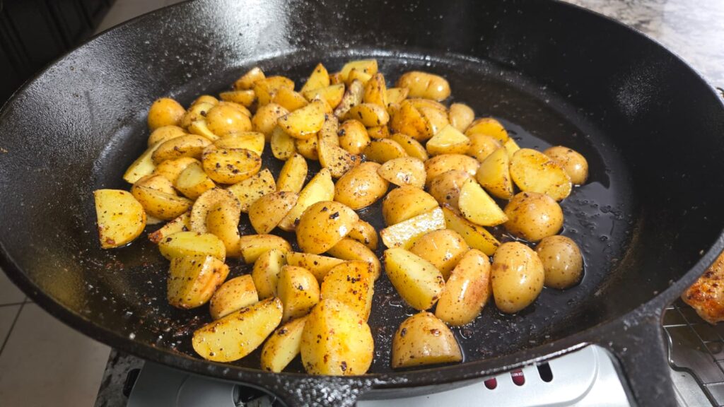 Small, golden potato pieces seasoned with herbs and spices are sizzling in a black cast iron skillet alongside succulent honey mustard chicken thighs, all cooking together on the stovetop.