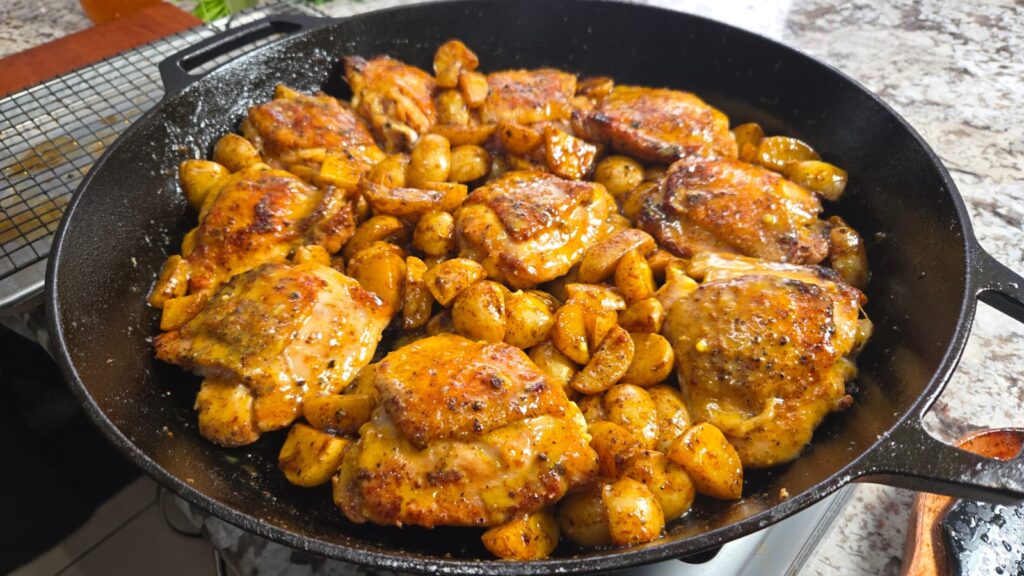 A cast-iron skillet filled with honey mustard chicken thighs and golden-brown baby potatoes sits seasoned and roasted to perfection on a kitchen counter.
