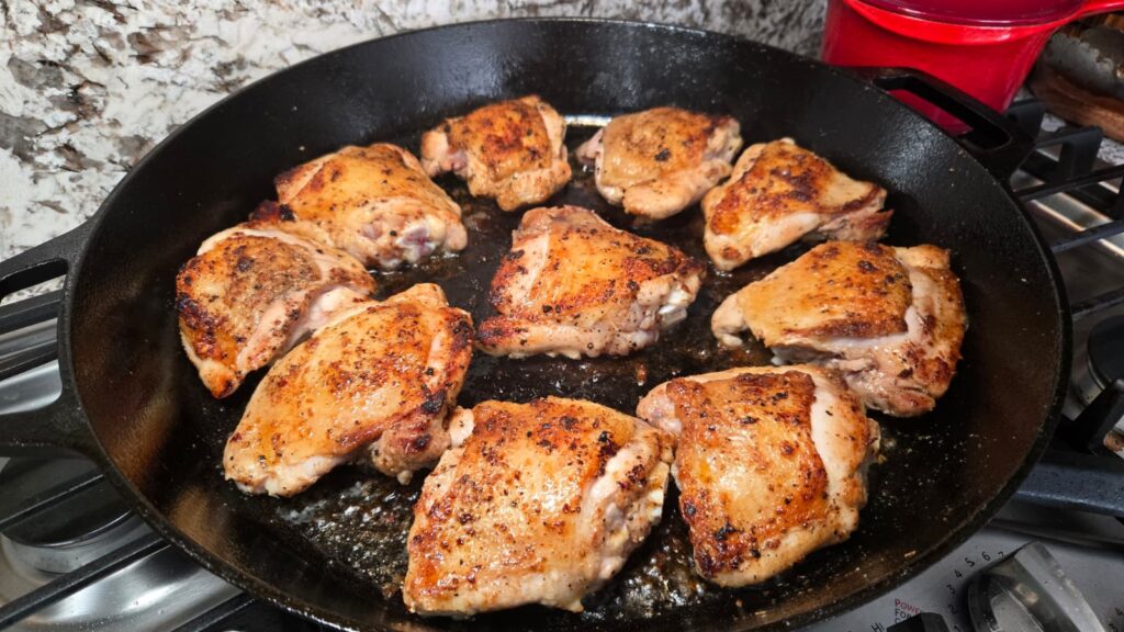 Ten browned honey mustard chicken thighs are cooking in a black cast iron skillet on a stovetop, with some oil visible at the bottom of the pan. The background shows a marbled surface and a red container.