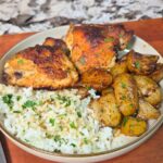 A plate with honey mustard chicken thighs, golden roasted potatoes, and white rice garnished with herbs. A signed chef’s knife rests beside the plate on a wooden board, with a fork and knife nearby.