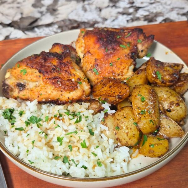 A plate with honey mustard chicken thighs, golden roasted potatoes, and white rice garnished with herbs. A signed chef’s knife rests beside the plate on a wooden board, with a fork and knife nearby.