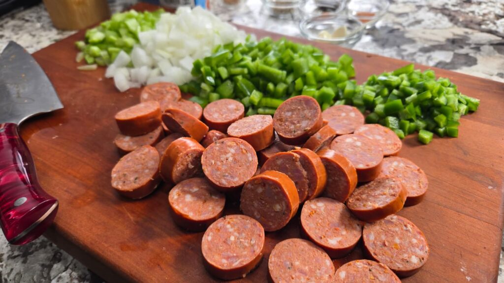 Sliced andouille sausage, chopped green bell pepper, celery, and onion are arranged on a wooden cutting board. A knife with a red handle rests nearby. The background shows a kitchen counter with additional ingredients for making andouille chili.