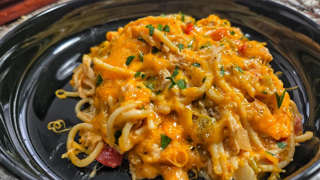 A close-up of chicken spaghetti baked to perfection, topped with melted cheddar cheese, shredded chicken, and chopped parsley, served in a black dish.