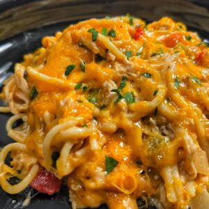 A close-up of chicken spaghetti baked to perfection, topped with melted cheddar cheese, shredded chicken, and chopped parsley, served in a black dish.