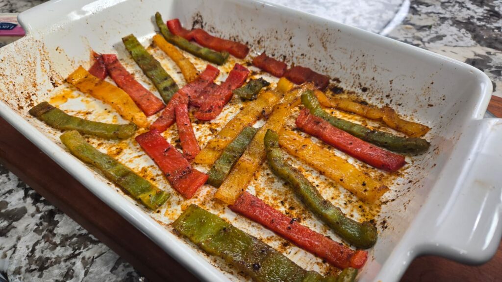 Roasted red, yellow, and green bell pepper strips are scattered in a white baking dish with seasoned residue left behind, set on a granite countertop.