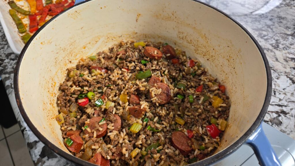 A pot of cooked rice mixed with sliced sausage, ground meat, green onions, and chopped red, yellow, and green bell peppers on a granite countertop.
