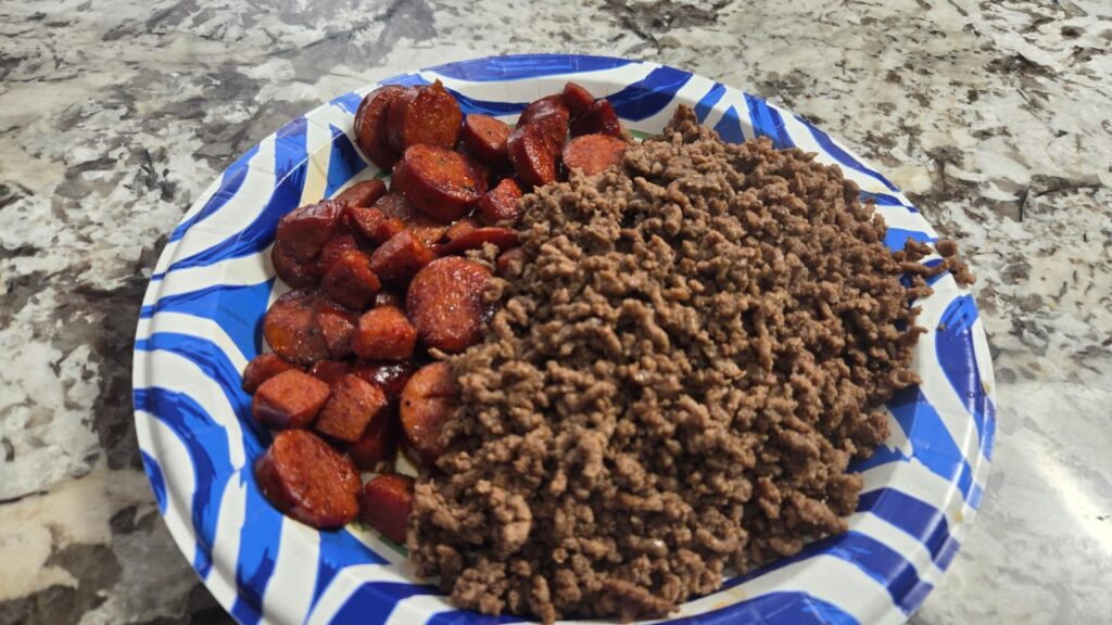 A blue and white paper plate on a marble countertop holds cooked ground beef and sliced, browned sausage pieces.