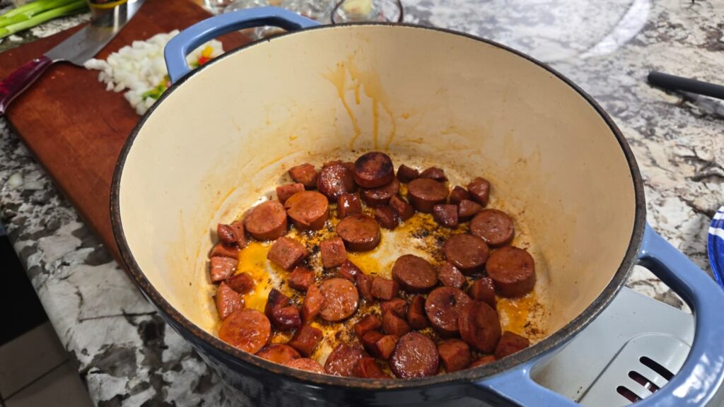 Sliced sausage browning in a large pot on a stovetop, with chopped vegetables and a knife on a wooden cutting board nearby.