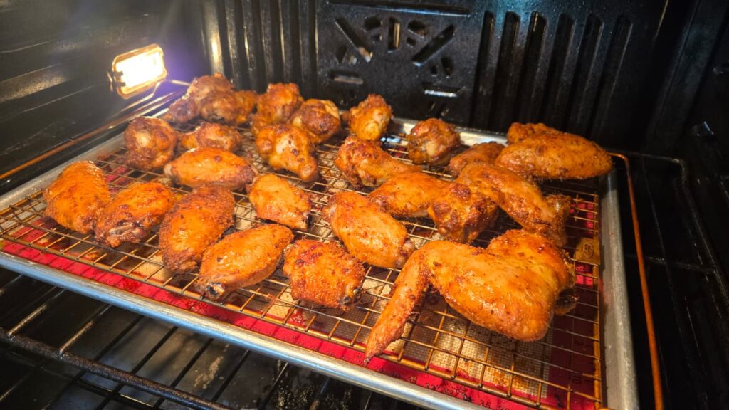 Chicken wings coated in seasoning are arranged on a wire rack over a baking sheet, being baked in an oven. The wings are golden brown and crispy, with the oven light illuminating the scene.