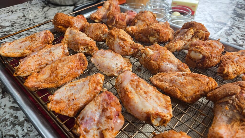 Raw chicken wings coated with seasoning are arranged on a wire rack over a baking tray, ready to be cooked. The background shows a marble countertop with jars of spices and other kitchen items.