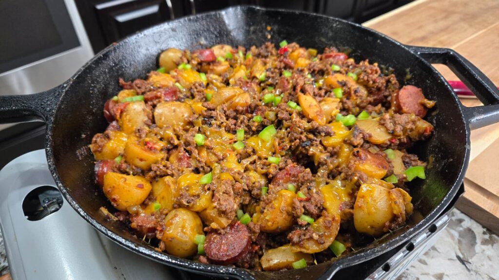 A cast iron skillet filled with dirty smothered potatoes, cooked ground meat, sliced sausage, melted cheese, halved potatoes, and chopped green bell peppers sits on a kitchen counter.