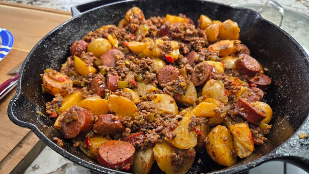 A cast iron skillet filled with dirty smothered potatoes, featuring cooked potatoes mixed with sliced sausage, ground meat, and diced vegetables, all seasoned and combined for a hearty dish.