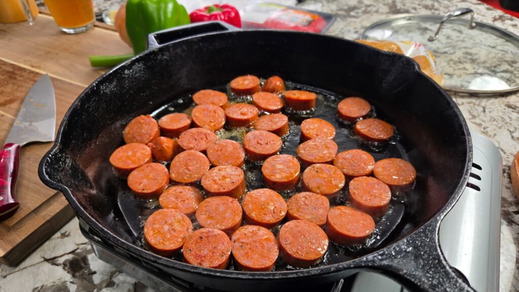 Sliced sausage cooking in a black cast iron skillet on a stove, with dirty smothered potatoes sizzling alongside vegetables and a knife on a wooden cutting board nearby.
