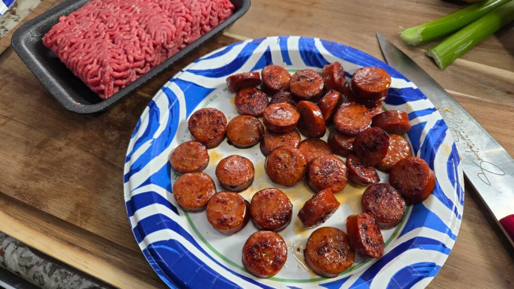 Sliced and cooked sausage on a blue and white paper plate, with raw ground beef, green onions, and a knife on a wooden cutting board&mdash;everything you need to start preparing hearty dirty smothered potatoes.