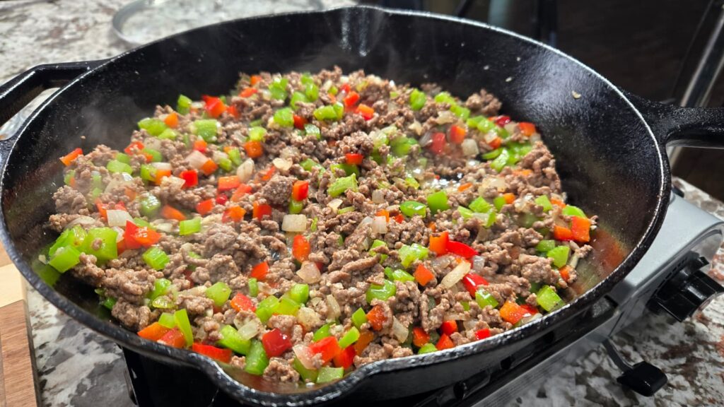 Ground beef, diced red and green bell peppers, onions, and dirty smothered potatoes cooking in a black cast iron skillet on a stovetop, with steam rising from the hearty mixture.
