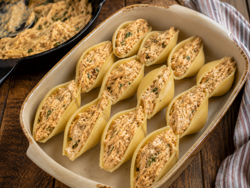 Large pasta shells filled with a seasoned, creamy chicken alfredo mixture are arranged in a baking dish, ready to be cooked. A skillet with more chicken alfredo stuffed shell filling is in the background, next to a striped kitchen towel on a wooden surface.