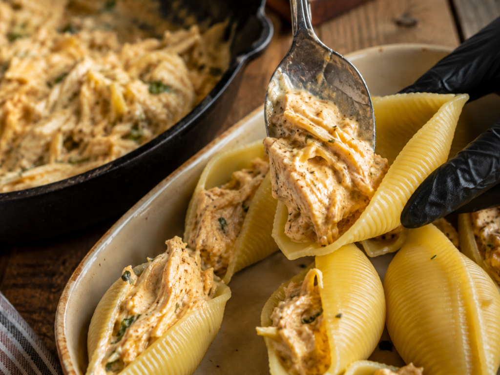 A gloved hand fills pasta shells with a creamy, seasoned chicken mixture from a pan, carefully preparing chicken alfredo stuffed shells on a rustic wooden table.