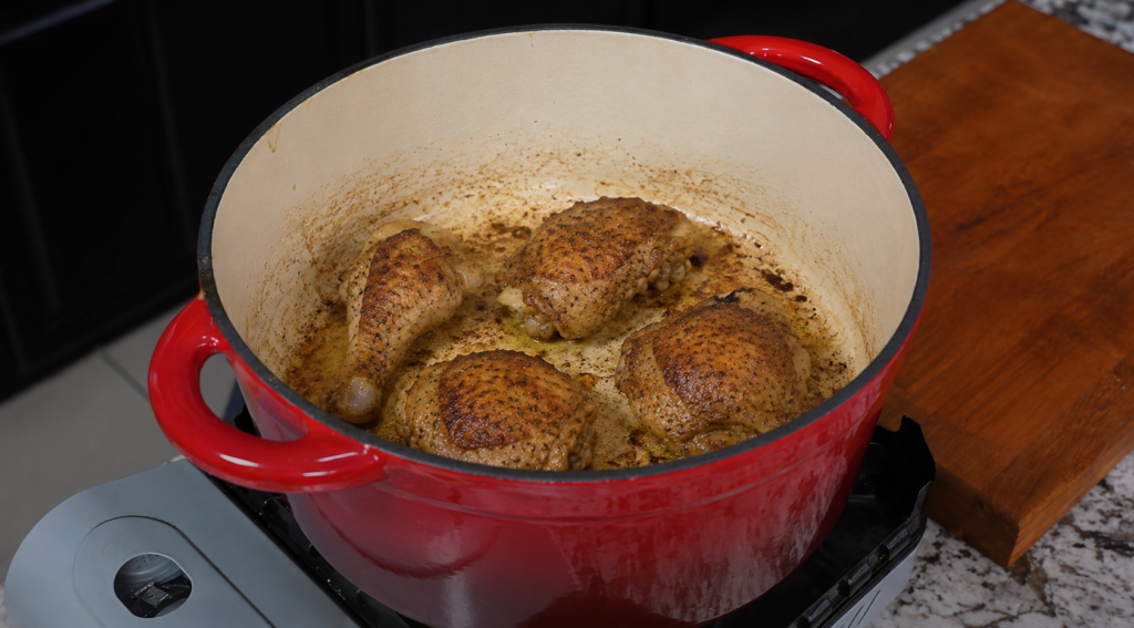 Four seasoned chicken pieces are browning in a red Dutch oven on a stovetop, with a wooden cutting board and granite countertop nearby.