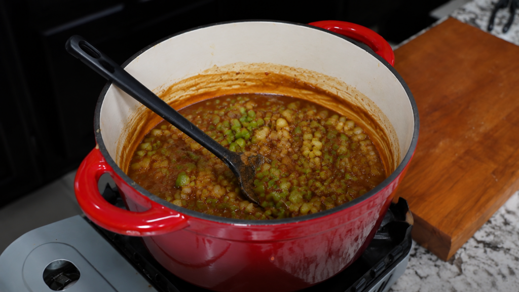 A red pot on a stove contains a thick, stew-like dish with green vegetables and beans, being stirred with a black spoon. A wooden cutting board sits nearby on a marble countertop.